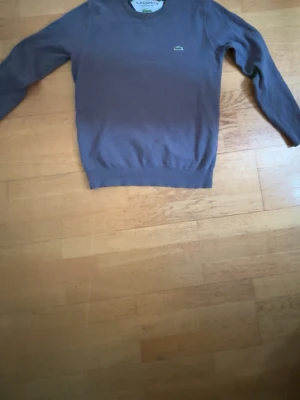 Grå tröja från Lacoste - Säljer en stilren grå tröja från Lacoste med den klassiska gröna krokodilloggan på bröstet. Tröjan har rund halsringning och långa ärmar. Perfekt för dig som gillar enkel och tidlös design. Inga defekter på tröjan. Hör av dig vid intresse eller frågor