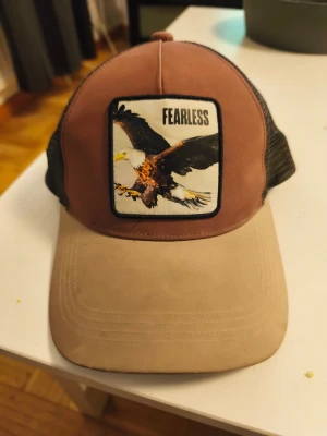 Brun och beige truckerkeps med örn - Säljer en brun och beige truckerkeps med svart mesh på sidorna och bak. Framtill finns ett stort tygmärke med en örn och texten 'FEARLESS'. Böjd skärm och justerbar passform. Perfekt för dig som vill sticka ut med en cool accessoar.