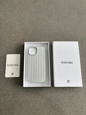 Silver mobilskal från Rimowa iPhone 15 - Elegant silver mobilskal från Rimowa med klassiska räfflade linjer och logga på baksidan. Skalet är tillverkat i tåligt material och har en stilren, modern design som ger din mobil en exklusiv look. Perfekt för dig som gillar minimalistisk och lyxig stil.