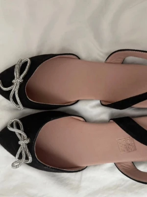 Svarta ballerinaskor med pärlrosett - Säljer ett par svarta ballerinaskor med spetsig tå och öppen sida. Skorna har en snygg dekoration framtill i form av en rosett av pärlor. Sula i gummi och insida i beige. Storlek 37. Perfekta för dig som gillar eleganta detaljer!