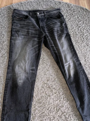 Svarta straight fit jeans från Diesel - Säljer ett par svarta straight fit jeans från Diesel med snyggt slitna detaljer och klassisk femficksmodell. Jeansen har en diskret logga på myntfickan och är tillverkade i mjuk denim med stretch för extra komfort. Perfekta för en cool och avslappnad stil.  W32. L32