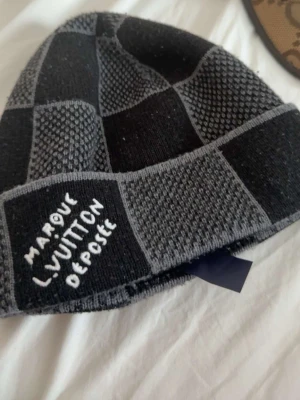 Gråsvart Louis Vuitton-mössa - Säljer en snygg grå och svart mössa från Louis Vuitton med rutigt mönster och texten 'MARQUE L.VUITTON DEPOSEE' broderad framtill. Mössan är stickad och har en modern, avslappnad passform. Perfekt accessoar för kalla dagar.