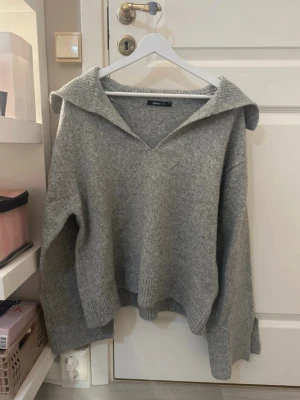 Grå stickad tröja med krage - Mysig grå stickad tröja från Gina Tricot med bred krage och v-ringning. Tröjan har långa ärmar, ribbade muddar och en avslappnad passform. Aldrig använd. Storlek M