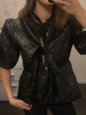 Svart glittrig blus med knyt - Säljer en svart glittrig blus med puffiga korta ärmar och flera knyt framtill. Blusen har en lätt strukturerad yta och en croppad känsla. Perfekt för dig som vill sticka ut med coola detaljer och trendig look. Pris går att diskutera 