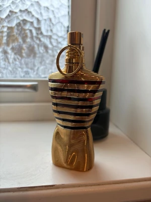 Jean Paul Gaultier Le Male parfym - Unik parfymflaska från Jean Paul Gaultier formad som en manlig torso i guld med svarta och röda ränder. Flaskan har en metallisk finish och en dekorativ ring runt halsen. Perfekt som statement-piece på parfymhyllan. 