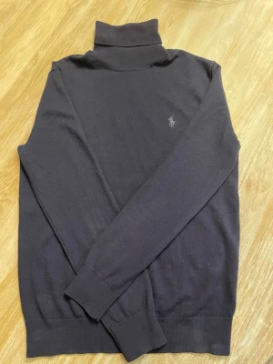 Svart polotröja från Polo Ralph Lauren - Klassisk svart polotröja från Polo Ralph Lauren i mjuk och tvättbar merinoull. Tröjan har lång ärm, ribbade muddar och diskret logga broderad på bröstet. Perfekt för lager på lager och en stilren look.