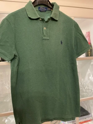 Grön pikétröja från Polo Ralph Lauren - Klassisk grön pikétröja från Polo Ralph Lauren i custom slim fit. Tröjan har korta ärmar, krage och två knappar framtill samt den ikoniska broderade loggan på bröstet. Perfekt för en clean och stilren look.