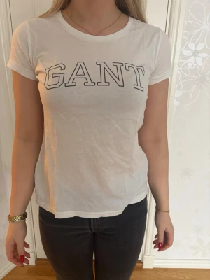 Vit GANT t-shirt med logga - Klassisk vit t-shirt från GANT med stor logga i svart på bröstet. T-shirten har rund halsringning och korta ärmar. Tillverkad i mjuk bomull som känns skön mot huden. Perfekt basplagg för en clean och enkel stil.