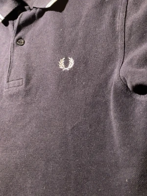 Mörkblå pikétröja från Fred Perry - Klassisk mörkblå pikétröja från Fred Perry med vit broderad logga på bröstet och knappar vid halsen. Tillverkad i 100% bomull för en skön och luftig känsla. Perfekt för en stilren och avslappnad look.
