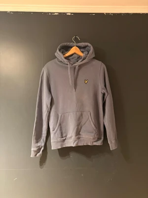 Grå hoodie från Lyle & Scott - Snygg grå hoodie från Lyle & Scott med klassisk gul logga på bröstet. Hoodien har huva med dragsko och en stor känguruficka framtill. Perfekt för en avslappnad och stilren look.