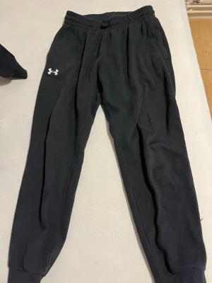 Svart loungewear-set från Under Armour - Mysigt svart loungewear-set från Under Armour med matchande hoodie och byxor. Hoodien har dragsko och magficka, medan byxorna har resår i midjan och muddar vid bensluten. Båda plaggen har Under Armour-logga framtill. Perfekt för chill dagar hemma.