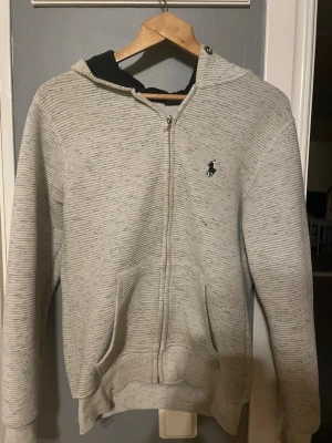 Gråmelerad hoodie från Polo Ralph Lauren - Säljer en gråmelerad hoodie från Polo Ralph Lauren med dragkedja framtill och två fickor. Klassisk logga broderad på bröstet och huva med svart innerfoder. Tillverkad i mjukt bomullsmaterial, perfekt för chill dagar eller när du vill ha en clean look.