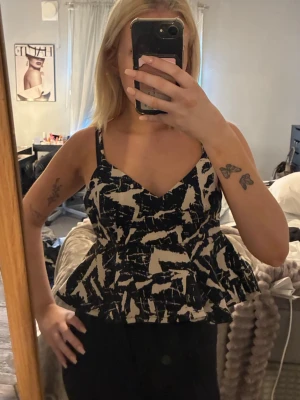 Svartvit  topp med mönster - Snygg peplum topp i svart och vitt med abstrakt mönster. Toppen har smala axelband, v-ringning och markerad midja som ger en feminin siluett. Perfekt att styla med jeans eller kjol för en trendig look.