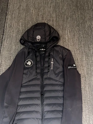 Svart Sail Antarctica Expedition-jacka  huva - Säljer en svart Sail Antarctica Expedition-jacka i fint skick. Jackan har en modern design med två tydliga patchar/loggor: 	•	Rund “Sail Antarctica Expedition”-patch på bröstet 	•	Rektangulär gummipatch på ärmen med Antarktis-motiv 	•	Koordinaterna “90°00’ S 00°00’ E” (Sydpolen) på båda patcharna