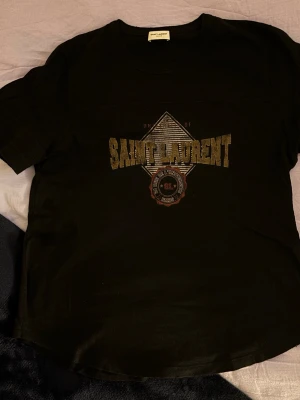 Svart Saint Laurent t-shirt - Säljer en svart t-shirt från Saint Laurent med stort tryck i guld och beige på bröstet. Klassisk rund halsringning och korta ärmar. Perfekt för dig som vill ha en stilren men ändå statement t-shirt från ett lyxigt märke.