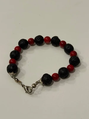 Armband med svarta lava pärlor och röda pärlor - Handgjort armband med svarta lavastenar och mindre röda pärlor. Armbandet har ett metallspänne i silverfärg som gör det enkelt att ta på och av.  