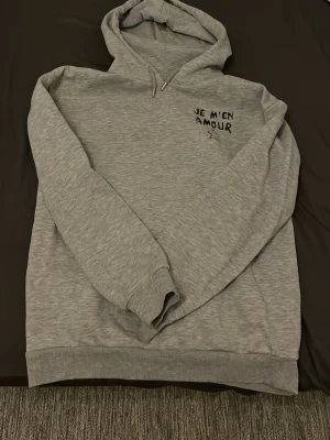 Grå hoodie med tryck 'JE M'EN AMOUR' - Säljer en ljusgrå hoodie med huva och dragsnören. På bröstet och ryggen finns svart tryck med texten 'JE M'EN AMOUR' och en stjärna. Hoodien har ribbade muddar vid ärmslut och nederkant. Perfekt för en avslappnad och trendig look.