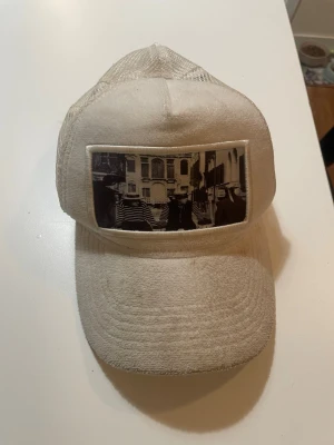 Beige keps från Emilio Pucci - Säljer en vit keps från Emilio Pucci med mesh på sidorna och baksidan för extra ventilation. Framtill finns en svartvit patch med ett grafiskt motiv. Kepsen har klassisk böjd skärm och justerbar passform.  