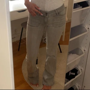 Grå bootcut jeans med fickdetaljer - Snygga grå jeans med bootcut passform och låg midja. Jeansen har breda framfickor och bakfickor med lock, vilket ger en cool och unik look. Tillverkade i klassiskt jeanstyg som är mjukt och följsamt. Perfekta för dig som gillar en avslappnad men trendig stil.
