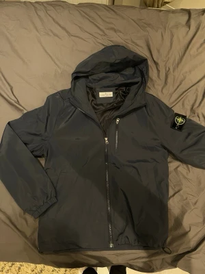 Marinblå vindjacka från Stone Island blå navy grisch archive  - Snygg svart vindjacka från Stone Island med huva och dragkedja framtill. Jackan har en ikonisk patch med logga på vänster ärm och en bröstficka med dragkedja. Tillverkad i lätt och slitstarkt material, perfekt för blåsiga dagar. Kom med bud, jag är lite desperat.