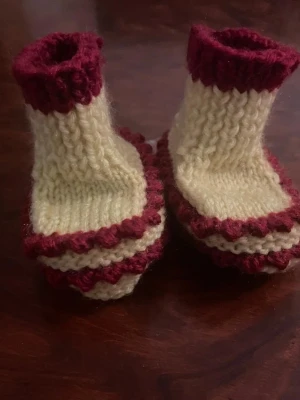 Stickade tofflor i vinrött och beige - Ny handstickade tofflor i mjukt garn med vinröda och beige ränder. De har en högre mudd upptill i vinrött och ett vågigt mönster längs kanterna. Perfekta för att hålla fötterna varma hemma och har en mysig retrostil.