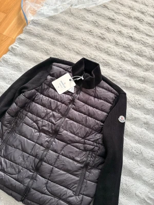 Moncler cardigan  - Snygg svart dunväst från Moncler med quiltad front och stickade svarta ärmar. Västen har dragkedja framtill, två sidofickor med dragkedja och Moncler-logga på ärmen. Materialet är en mix av dun och stickat tyg för extra komfort och stil.