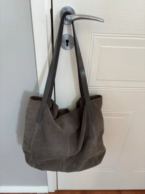 Grå mocka shoppingväska - Stilren shoppingväska i grå mocka med långa, mörkgrå axelband i slätt läder. Väskan har en mjuk och rymlig form, perfekt för att bära med sig allt man behöver. Inga synliga loggor eller märken på utsidan.