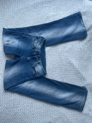Blå bootcut jeans med slitningar - Säljer ett par blå bootcut jeans med snygga slitningar och tvättade detaljer. Jeansen har klassisk femficksdesign, knappgylf och raka ben. Perfekta för en avslappnad och trendig look.