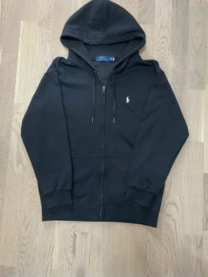 Polo Ralph Lauren Zip Hoodie – Stilren & eftertraktad 🔥 - Snygg och populär Polo Ralph Lauren zip-hoodie i klassisk svart färg. Ett perfekt basplagg som kombinerar komfort och premiumkänsla. Lätt att matcha och passar lika bra till vardag som till en mer uppklädd streetwear-look.  • Märke: Polo Ralph Lauren   • Typ: Zip hoodie / huvtröja med dragkedja   • Färg: Svart   • Passform: S kanske Xs • Skick: Väldigt bra skick   • Detaljer: Huva med snören, hel dragkedja, muddar vid ärmslut och nederkant, fickor fram • Mjuk och skön kvalité! 