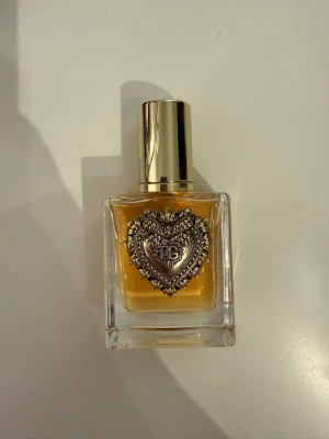 Dolce & Gabbana Devotion EdP 50ml - Lyxig parfym från Dolce & Gabbana i en elegant, fyrkantig glasflaska med guldfärgat hjärtdetalj framtill och guldfärgad kork. Flaskan innehåller 50 ml Eau de Parfum med bärnstensfärgad vätska. Perfekt för dig som gillar exklusiva dofter och snygg design.
