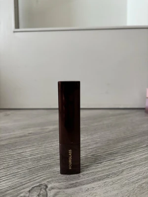 Hourglass foundation stick beige - Hourglass foundation stick i en elegant mörkbrun hylsa med guldtext. Produkten har en krämig konsistens och kommer i en beige nyans. Den trekantiga formen gör det enkelt att applicera foundationen jämnt i ansiktet.