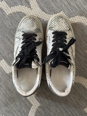 Sneakers från Zadig & Voltaire, ormmönstrade - Säljer ett par vita sneakers från Zadig & Voltaire med svart ormmönster och svarta skosnören. Skorna har en cool blixtdetalj på sidan och silverfärgad häl. Använda fåtals gången då storleken är fel. 
