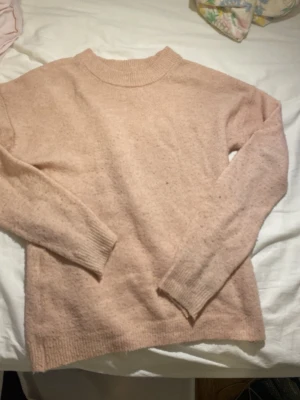 Ljusrosa stickad tröja - Mysig ljusrosa stickad tröja med rund halsringning och långa ärmar. Tröjan har ribbstickade muddar vid ärmslut och nederkant, vilket ger en klassisk och enkel look. Perfekt för kyliga dagar och lätt att matcha med olika outfits.