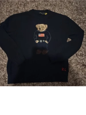 Mörkblå stickad tröja med björnmotiv - Säljer en mörkblå stickad tröja från Ralph Lauren med det klassiska björnmotivet på bröstet. Tröjan har långa ärmar, rund halsringning och RL-broderi nertill. Perfekt för dig som gillar preppy stil och detaljer.