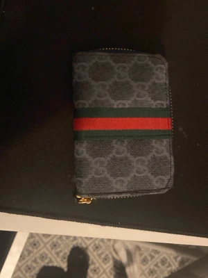 Gucci plånbok med monogram och ränder - Snygg plånbok från Gucci i mörkgrått tyg med klassiskt GG-monogram. Plånboken har gröna och röda ränder på framsidan och dragkedja i guld. Perfekt för dig som gillar lyxiga accessoarer med ikonisk design.