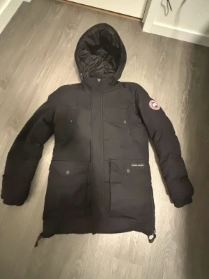 Svart dunjacka från Canada Goose - Snygg svart dunjacka från Canada Goose med huva och klassisk logga på ärmen. Jackan har flera praktiska fickor med knappar och dragkedja framtill. Perfekt för kalla vinterdagar och riktigt varm tack vare dunfyllningen.