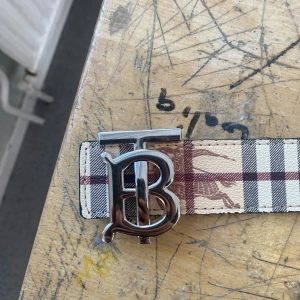 Burberry bälte med TB-spänne - Snyggt Burberry-bälte med klassiskt rutigt mönster i beige, brun, svart och vit. Bältet har ett stort metallspänne med TB-logga och är gjort i skinn. Perfekt accessoar för att lyfta din outfit.