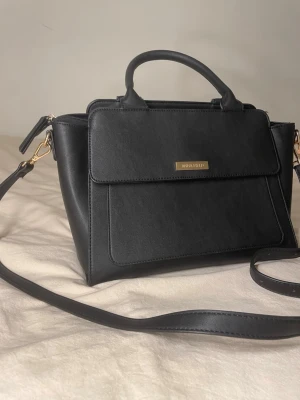 Svart handväska från Anna Field - Stilren svart handväska från Anna Field i slätt konstläder med guldfärgade detaljer. Väskan har både handtag och avtagbar axelrem, samt en praktisk ficka framtill med lock. Perfekt för dig som gillar klassisk och enkel design.
