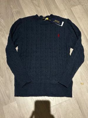 Mörkblå kabelstickad tröja Polo Ralph Lauren - Kabelstickad mörkblå tröja från Polo Ralph Lauren med rund halsringning och röd broderad logga på bröstet. Långa ärmar och ribbade muddar ger en klassisk look. Perfekt för lager-på-lager-stil.