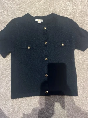 marinblå kortärmad kofta Gina Tricot - Kortärmad marin blå kofta från Gina Tricot med guldfärgade knappar framtill och dekorativa fickor på bröstet. Ribbstickade detaljer vid halsringning, knappslå och ärmslut. Perfekt för att lyfta en enkel outfit.