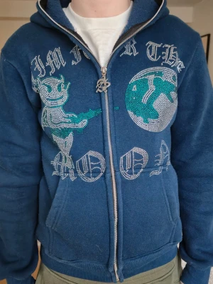 Blå hoodie från Aim For The Moon - Säljer en blå hoodie med dragkedja från Aim For The Moon. Den har huva, fickor och ett stort tryck med text och motiv av en figur och måne i silver och turkos rhinestones på framsidan. Perfekt för dig som gillar unika och coola plagg. Köpt direkt från Aim for the Moon. 