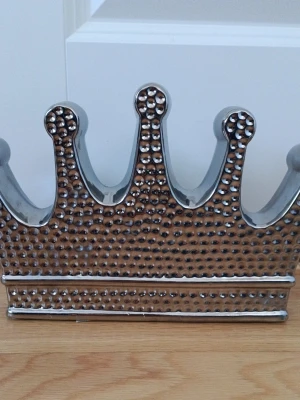 Silverfärgad prydnads krona i keramik - Dekorativ krona i blank silverfärgad keramik med prickig struktur på framsidan. Kronan har en klassisk form med fem toppar och är ihålig, perfekt som prydnad på hyllan eller fönsterbrädan. Ger ett lyxigt och stilrent intryck i rummet. Skriv till mig på Plick om ni vill ha måt på den.