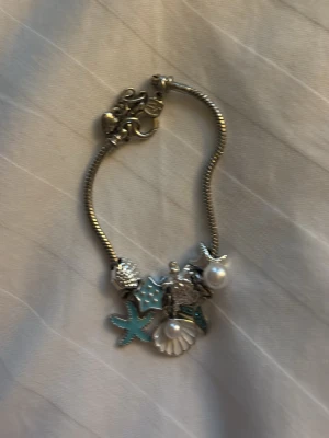 Silverfärgat armband med havstema - Säljer ett snyggt silverfärgat armband med marina berlocker som sjöstjärna, snäcka, sköldpadda och pärlor. Några detaljer är turkosa och armbandet har en smidig låsning. Perfekt accessoar för dig som gillar strandvibbar och unika smycken.