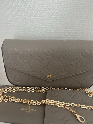 Beige Louis Vuitton kuvertväska - Säljer en beige kuvertväska från Louis Vuitton i präglat läder med det klassiska monogrammönstret. Väskan har en elegant, avlång form och stängs med en guldfärgad knapp framtill. Perfekt accessoar för dig som gillar lyxiga detaljer och stilren design.