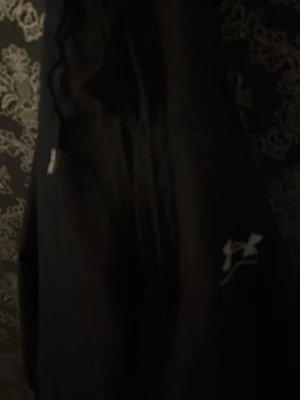 Svart träningsjacka från Under Armour - Säljer en svart träningsjacka från Under Armour med dragkedja och logga på ärmen. Jackan har sportig design och är tillverkad i ett lätt, funktionellt material som passar perfekt till träning och aktivitet.