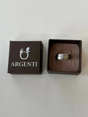 Silverring från Argenti - Snygg silverfärgad ring från Argenti med ett diskret, ingraverat bladliknande mönster runt om. Ringen har en bred, klassisk form och är tillverkad i metall med blank finish. Perfekt för dig som gillar stilrena accessoarer med en twist. Den är justerbar och handtillverkad. 