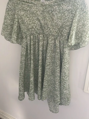 Två somriga kortklänningar - Säljer två korta klänningar, en grönblommig med omlott och volangärmar samt en vit med små guldprickar och volanger. Båda har luftig passform och är perfekta för varma dagar.