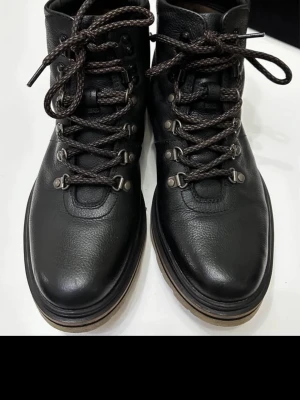 Klarks Svarta läderkängor med grova snören - Säljer ett par svarta kängor i äkta läder med grova, mörkbruna snören och metallhakar. Kängorna har en robust sula i kontrastfärg och en klassisk outdoor-look. Perfekta för dig som gillar stilrena och funktionella skor.