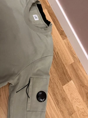 Ljusgrön sweatshirt från C.P. Company - Snygg ljusgrön sweatshirt från C.P. Company med rund halsringning och ribbade muddar. Tröjan har en unik detalj med ficka och svart lins på ärmen, vilket ger en cool streetwear-vibe. Tillverkad i mjuk bomull för skön komfort.