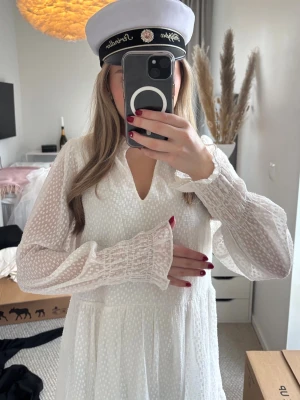 Vit klänning med volang - Söt vit kortklänning med volangdetaljer. Klänningen har långärmade, lätt transparenta ärmar med struktur och knyt i midjan. Perfekt för student eller sommarens tillfällen. Helt oanvänd med prislapp kvar. 